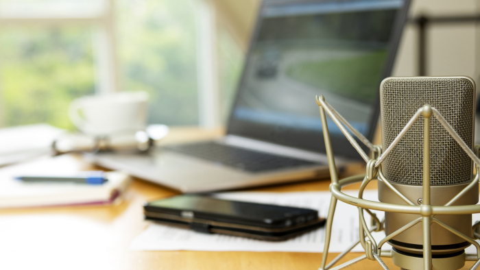 A desk set up with a microphone in the foreground and a laptop, phone, paper and pencil, glasses, and a mug