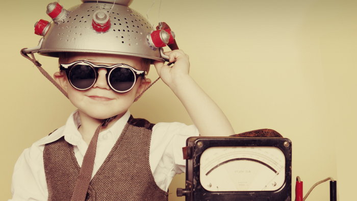 A young boy wearing goggles with a metal colander on his head sitting next to a machine 