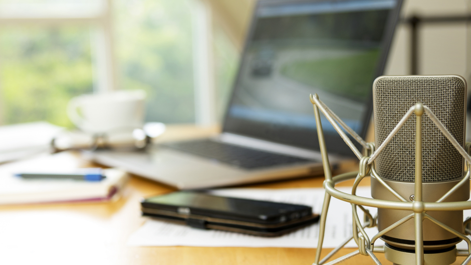 A desk set up with a microphone in the foreground and a laptop, phone, paper and pencil, glasses, and a mug